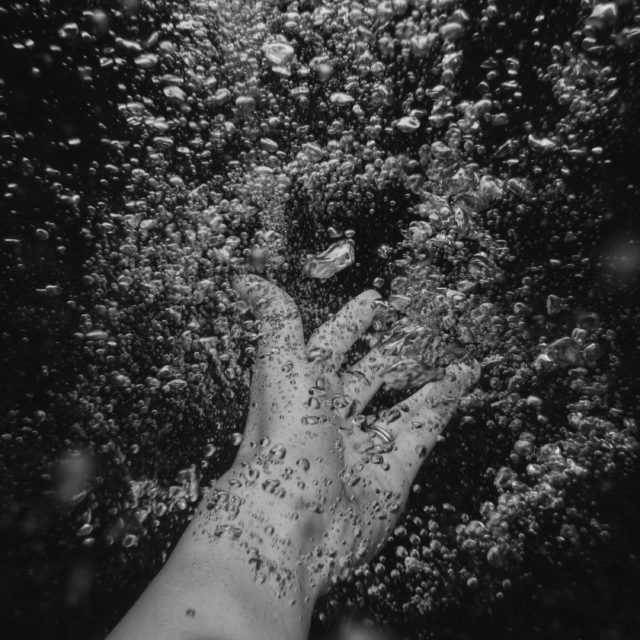 A black and white image of a hand reaching out underwater, surrounded by bubbles