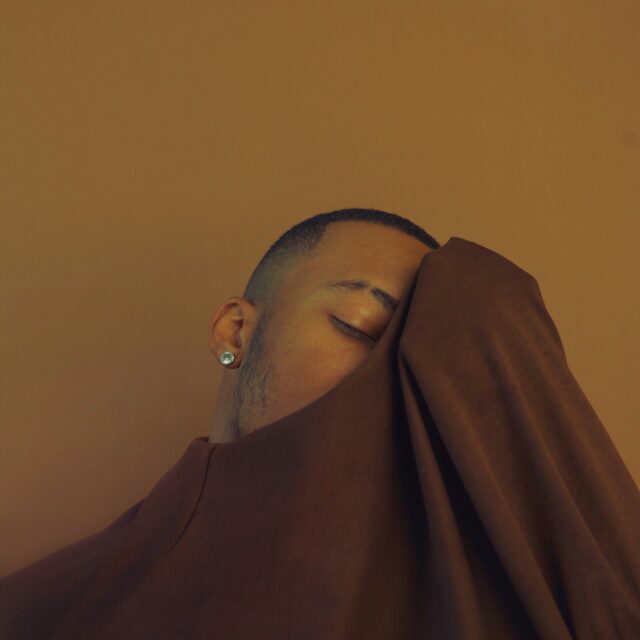 A person of colour holds a brown shirt up over half of their face