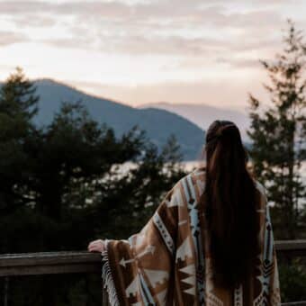 A woman in a poncho stares out at mountains, pines and a river
