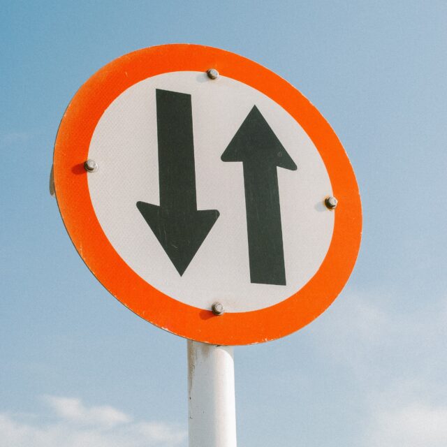 Opposing arrows in a red circle on a road sign. The background is the sky.
