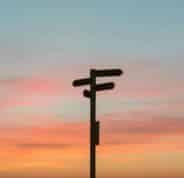 A directional sign post pointing in multiple directions silhouetted against a sunset with colours of burning orange and blue.