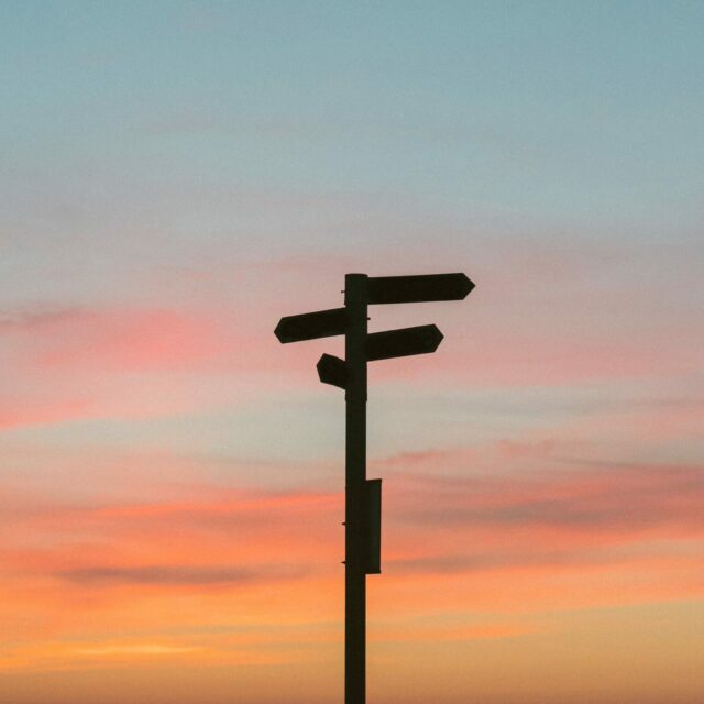 A directional sign post pointing in multiple directions silhouetted against a sunset with colours of burning orange and blue.
