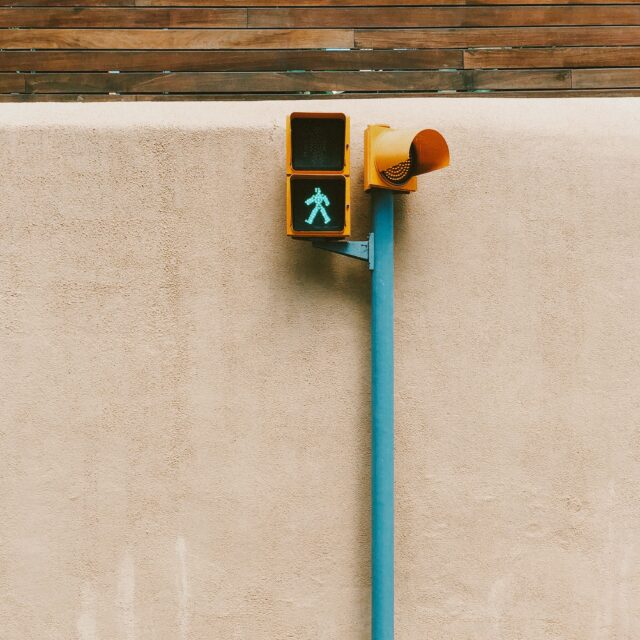 A single cross walk signal sign lit up with the green walking sign. The sign is yellow with a green post and it stands in front of a concrete wall with wood panelling above.