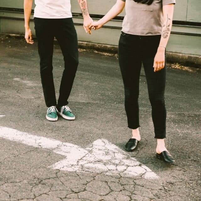 Two people stand in front of a double ended arrow painted on the pavement. One person is guiding the other in one direction of the arrow by holding their hand.