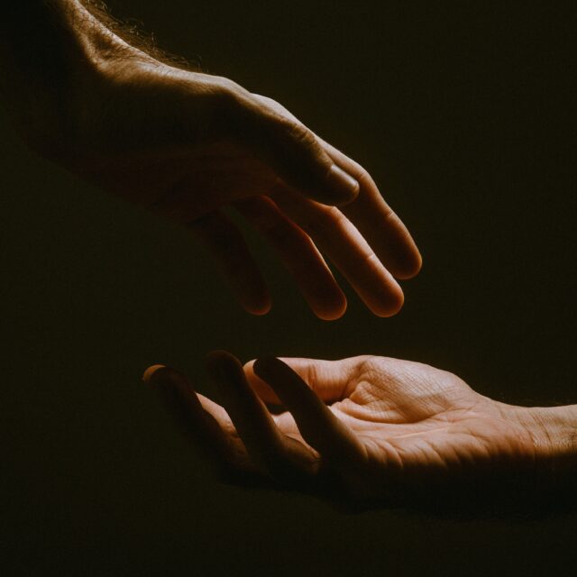 Two hands reach for each other gently lit by one light casting shadows on the hands.