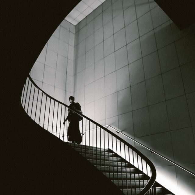 A silhouette is seen climbing up a large winding stair case that is it by light on the edge of the stairs. The walls are tiled in a minimalist manner.