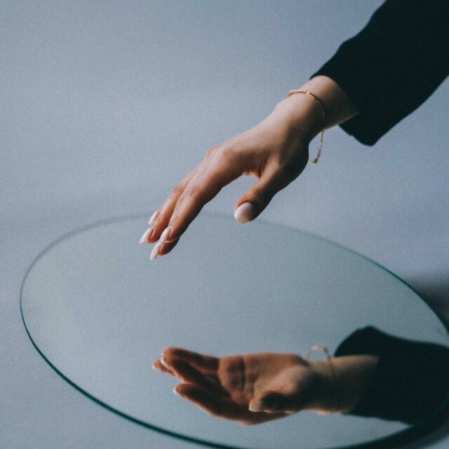 A hand is seen reaching towards a circular mirror. The hand is reflected, the backdrop is plain and white.