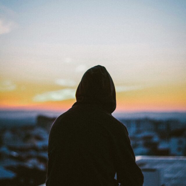 A hooded silhouette sits on the roof of a building observing the sunrise of a city.