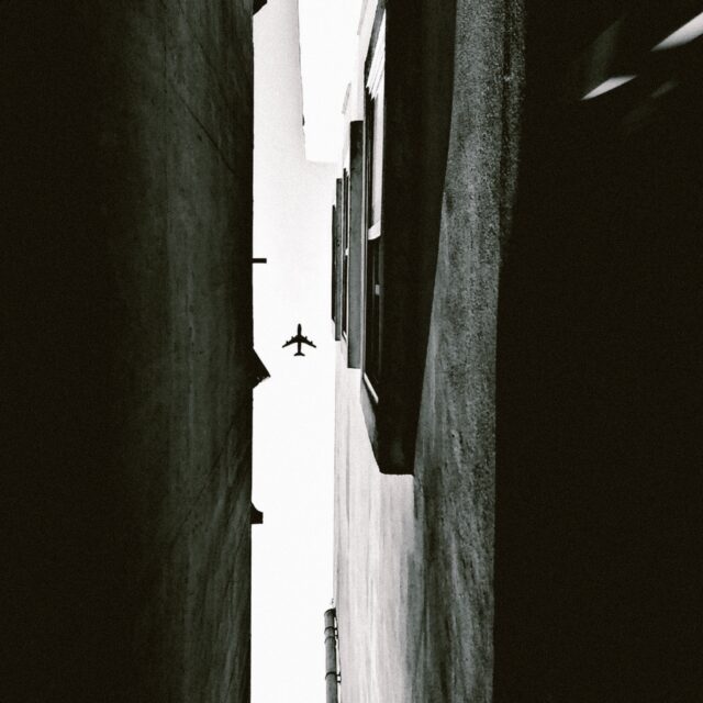 A worms eye view looking up from a small alley way. The space of sky between two buildings is seen, a plane is captured in the sky.