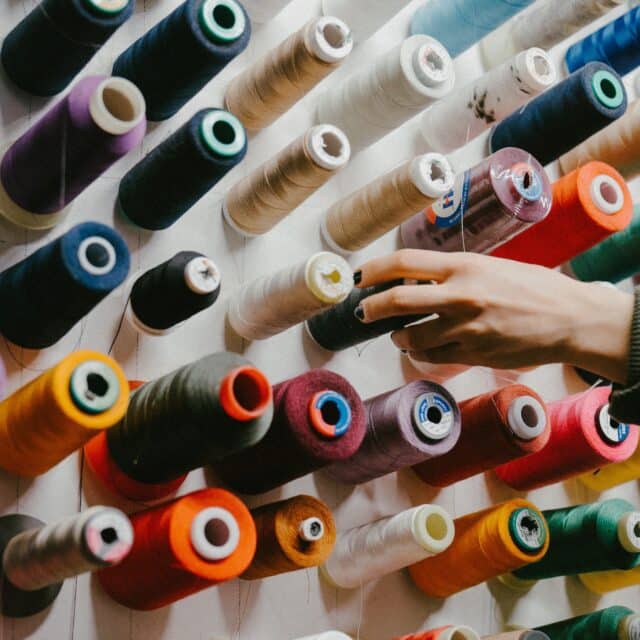 Image of a hand reaching for a bob of thread on a wall. It is surrounded my multi-coloured thread bobs all mounted to a wall.