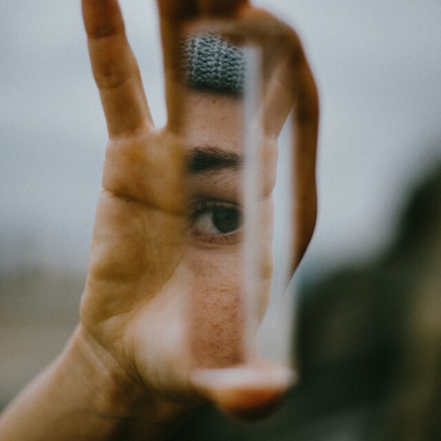 An eye is seen reflected and reflected through a glass prison which is held by a hand. The background is blurred.