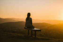 A person sits on a bench that overlooks a valley. The sun is setting providing a warm glow from the right of the image.