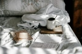 A white coffee mug sits atop a bed next to a journal